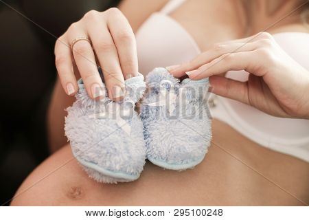 Pregnant Woman Holding Pair Of Blue Shoes For Baby Boy. Daylight. Indoor Shot