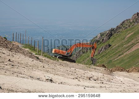 An Orange Digger Digging A Pit On Top Of The Mountain Overlooking The City. Photo Taken In Kazakhsta