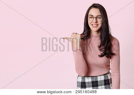 Positive Smiling Slender Model Standing Over Light Pink Background, Showing Direction With Her Thumb