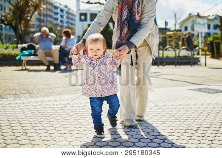 Grandmother Helping Granddaughter Make Her Firts Steps. Middle Aged Woman With Little Girl On The Se