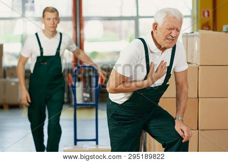 Senior Warehouseman Having A Heart Attack At Work, Young Colleague Running To Help Him