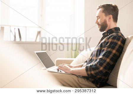 Young casual man pushing keys of laptop keypad while sitting on couch in room and carrying out remote work