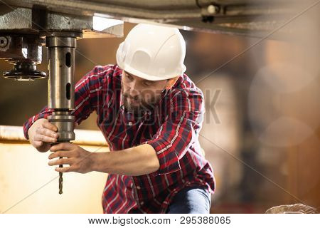 Young technician in hardhat and overalls putting drill element into iron holder which is part of large industrial machine