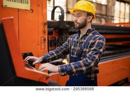 Young contemporary engineer in uniform and hardhat standing by terminal while controling industrial machine
