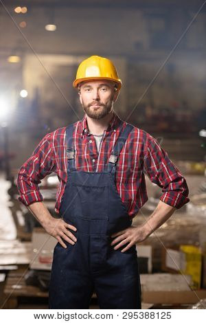 Young confident machinebuilder in workwear keeping his hands on waist while standing right in front of camera inside workshop