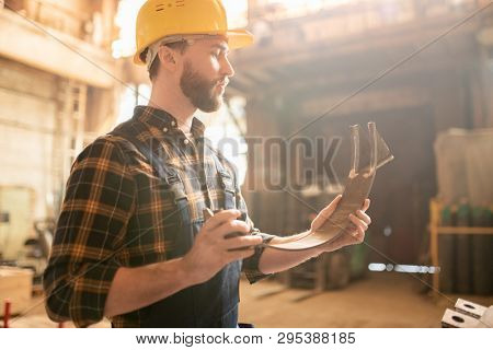 Young man in overalls and hardhat looking at metallic semi-circle detail in his hands while standing in workshop