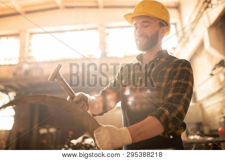 Young worker of contemporary machinebuilding plant holding metallic workpiece and hammering it
