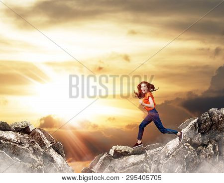 Redhead Woman Jumps Through The Gap Between Hills Over Cliff On Blue Sky Background.