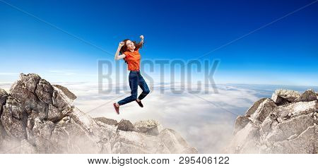 Redhead Woman Jumps Through The Gap Between Hills Over Cliff On Blue Sky Background.