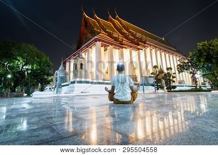 Man Meditating In Lotus Pose In Wat Suthat Thepwararam At Night, Reflection In The Floor Marble Tile