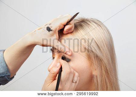 Visagiste With A Brush In One Hand Putting Makeup On The Eyes Of The Model, The Other Hand Pulls The
