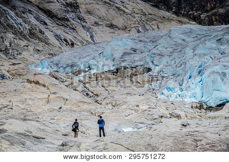 Close View Of Nigardsbreen Glacier In Jostedal With Two Tourists Looking At The Glacier