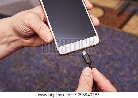 Female Hand Inserts The Usb Cable Charger Into The Mobile Phone Against The Background Of The Apartm
