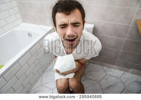 A Young Guy Sits On The Toilet And Holds Toilet Paper. He Is In The Bathroom And He Feels Bad