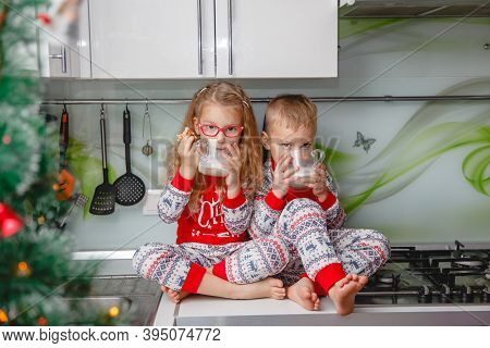 Little Brother And Sister Sit On A Christmas-decorated Kitchen Table In Pajamas And Drink Milk. Girl