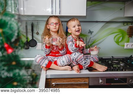 Little Brother And Sister Sit On A Christmas-decorated Kitchen Table In Pajamas And Drink Milk. Girl