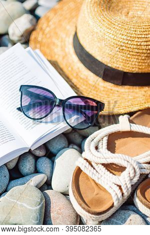 Beach Hat On Opened Book With Sunscreen And Shoes On Pebble Beach