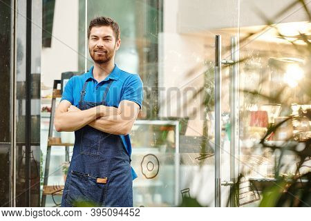 Portrait Of Smiling Confident Cafe Waiter Stading At Coffeshop Entrance With Arms Folded And Looking