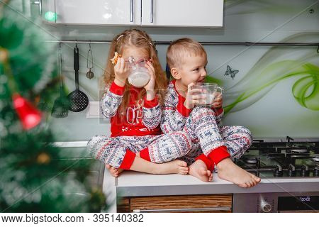 Little Brother And Sister Sit On A Christmas-decorated Kitchen Table In Pajamas And Drink Milk. Girl