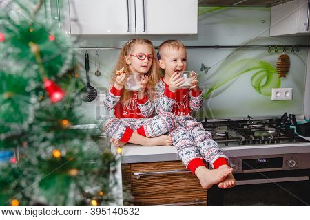 Little Brother And Sister Sit On A Christmas-decorated Kitchen Table In Pajamas And Drink Milk. Girl