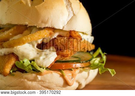 Close Up Of A Homemade Chicken Burger With Onion Rings On A Chopping Wood Board, On Black Background