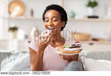 Young Black Woman On Diet Having Cheat Meal Day, Holding Plate Of Sweets, Stuffing Her Mouth With Do