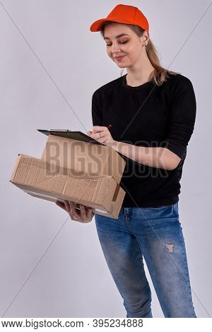 Smiling Female Courier Is Signing Documents. European Girl In Orange Cap Is Holding Parcels. Isolate
