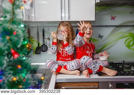 Little Brother And Sister Sit On A Christmas-decorated Kitchen Table In Pajamas And Drink Milk. Girl