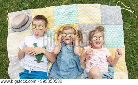 Kids Smile And Indulge Lying On A Patchwork Rug. Children Fooling Around With A Cucumber Mask. The B