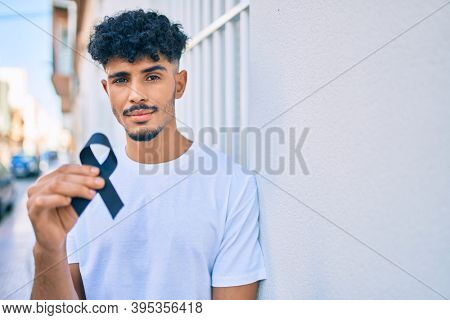Young arab man with serious expression holding mouring black ribbon leaning on the wall.