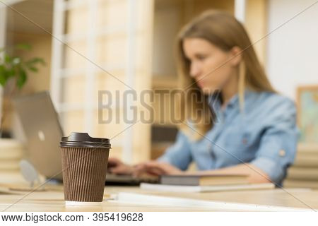 Selective Focus On A Coffee Paper Cup On The Table Young Woman Working On Her Computer On The Backgr