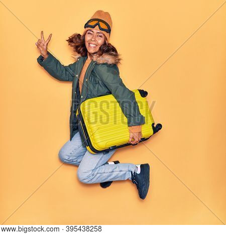 Young beautiful latin woman wearing winter clothes smiling happy. Jumping with smile on face holding cabin bag doing victory sign over isolated yellow background