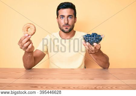 Young hispanic man eating breakfast holding donut and blueberries relaxed with serious expression on face. simple and natural looking at the camera. 