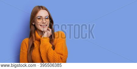 Ginger Woman With Red Hair Is Smiling At Camera With Glasses On A Blue Studio Wall With Free Space