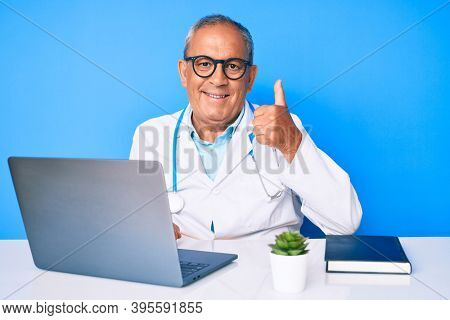 Senior handsome man with gray hair wearing doctor uniform working using computer laptop doing happy thumbs up gesture with hand. approving expression looking at the camera showing success. 