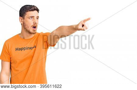 Handsome young man with bear wearing tshirt with happiness word message pointing with finger surprised ahead, open mouth amazed expression, something on the front 