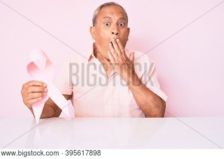 Senior handsome man with gray hair holding pink cancer ribbon sitting on the table covering mouth with hand, shocked and afraid for mistake. surprised expression 