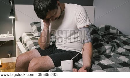 Young Caucasian Man Measures Blood Pressure At Home In Bedroom Sitting On Bed At Home Clothes. Male 