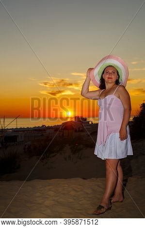Woman In A White Dress And A Straw Hat On A Background Of The Sea At Sunset.