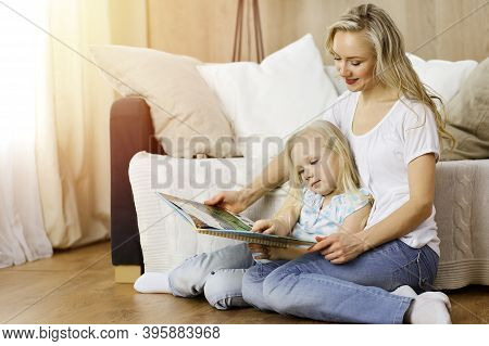 Happy Family. Blonde Young Mother Reading A Book To Her Cute Daughter While Sitting At Wooden Floor 