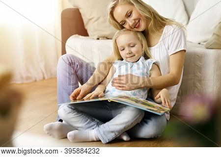 Happy Family. Blonde Young Mother Reading A Book To Her Cute Daughter While Sitting At Wooden Floor 