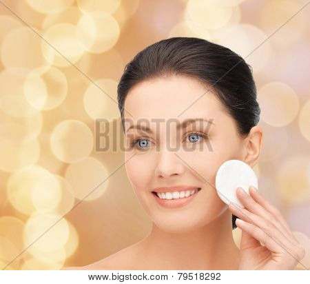 beauty, people and health concept - beautiful smiling woman cleaning face skin with cotton pad over beige lights backgroundd