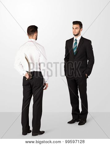 Two opposing businessman shake hands, one of them hiding a weapon behind his back
