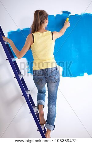 Happy beautiful young woman doing wall painting, standing near ladder