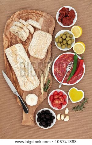 Italian antipasti selection with olives, salami, chilli peppers, sun dried and fresh tomatoes, garlic, lemon, oil, with ciabatta bread on an olive wood board on brown ridged paper  background.