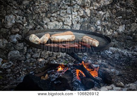 Sausages and buns being grilled on a ganging grill in front of a stone wall