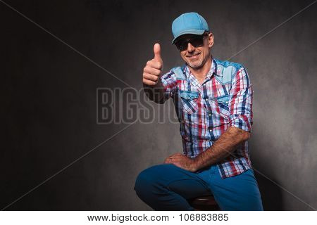 positive old casual man in trucker hat making the ok thumbs up hand sign while sitting in studio
