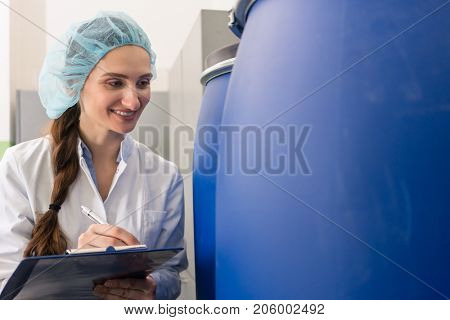 Motivated manufacturing employee writing a report while checking the plastic containers, with liquid cosmetics during quality control in the factory