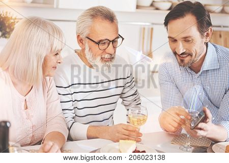 Have a look here. Cheerful adult man showing his parents smartphone while sitting at the table and enjoying meal together