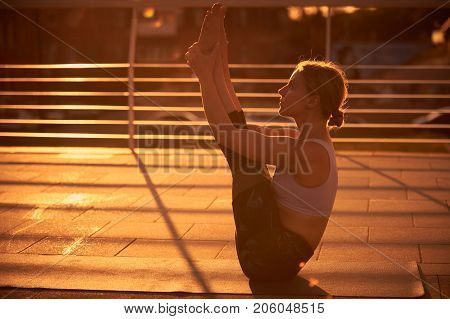 Young attractive woman practicing yoga, sitting in Paripurna Navasana exercise in the terrace at sunset